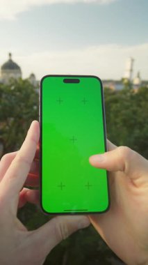 Yeşil ekranı gösteren telefonları olan yakın plan erkek elleri. Genç bir adam akşam parkında yürürken arkadaşıyla mesajlaşıyor. Arka planda eski şehir mimarisi, dışarıda. Yaz zamanı. Krom anahtar kavramı. Dışarıda.