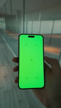 Yeşil ekranlı, el ele tutuşan ve dikey olarak telefonu kaydıran adamı kapat. Arka planda büyük pencereli modern alan. Akıllı telefondan yazıyorum.