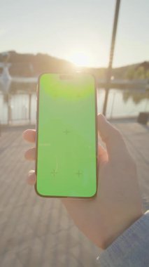 Yeşil ekranlı, dikey telefon akıllı telefon cihazını tutan eller hareket ediyor. Arka plandaki gölde, güneş ışığında. Doğa
