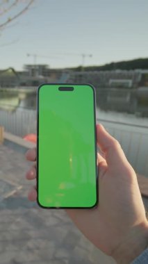 Yeşil ekran yürüyüşlü, dikey telefon cihazını tutan genç elleri kapat. Arka plandaki gölde, güneş ışığında. Doğa