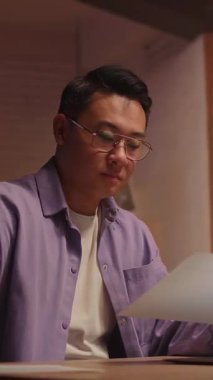 Gözlüklü Asyalı iş adamı dizüstü bilgisayar kullanıyor ve telefonda konuşuyor. Geceleri ekrana ciddi ciddi bak. Kağıt üzerinde çalışmak. 360 derece. İz sürme görüntüsü etrafında hareket