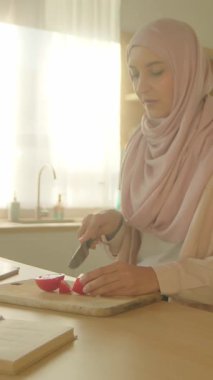 Günışığında tesettürlü müslüman kadın masada oturmuş modern ışık mutfağında domates kesiyor. Sebze. Kamera hareket ediyor. 360 derece