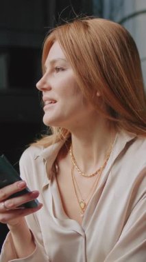 Kızıl saçlı, zarif kıyafetli, gülümseyen genç bir kadının portresi modern otelin koridoruna bakıyor. Akıllı telefonu tutan ve içeride sohbet eden çekici bir kız.