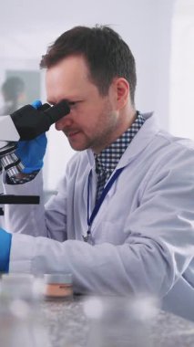 Beyaz labirent giymiş genç beyaz bir adamın mikroskopla laboratuvara baktığı görüntüler. Mikroskopta mikrobiyolojik madde inceleyen ve laboratuvar kavanozu kullanan bir adam. Bilim adamı çalışıyor. Kapalı
