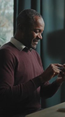 Rahatlamış gülümseyen Afrikalı adamın portresi akıllı telefonu elinde tutuyor, ekrana bakıyor, sohbet ediyor. Çalışanların işten izin alıp eğlendiği bir fotoğraf. Modern çatı katında akıllı telefon kullanan serbest çalışan.