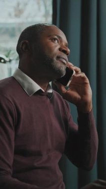 Sinirli bir Afrikalı adamın bilgisayarın önünde oturup akıllı telefondan zor bir konuşma yapması. İş adamı aktif olarak el kol hareketi yapıyor, iş ortağıyla konuşuyor. İçeride. Modern iç mimari