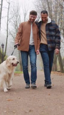 Gülümseyen, aynı cinsiyetten erkek, çok ırklı bir çift köpekle parkta yürüyor.