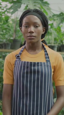 Serada önlük giyen, kollarını kavuşturup kameraya poz veren Afro-Amerikan profesyonel kadın çalışanın portresi. Tarım. Organik gıda ekimi.