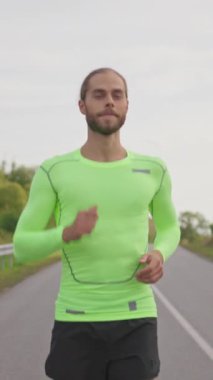 Yeşil, parlak spor kıyafetli, kır yolunda koşan, gülümseyen koşucu yakışıklı adam. Atletik adam sabah antrenmanı yapıyor. Kırsal otoyol.