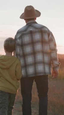 Kafkasyalı yetişkin bir baba, sevimli oğluyla el ele tutuşup dışarıda buğday tarlası köyünde yürüyor. Yaz batımı. Manzara. Aile kavramı.