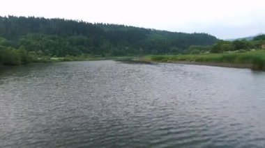 Fevkalade sakin bir nehir manzarası bulutlu bir gökyüzünün altında uzanan yemyeşil tepelerin arasında yuva yapar.