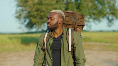 Sırt çantasıyla gezen ve kameraya bakan yakışıklı Afro-Amerikan adam yaz günü güzel Afrika bozkırlarında kalıyor. Safari kavramı.