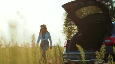 Şapkasıyla arabasının yanında yürüyen mutlu ve çekici genç bir kadın. Gün batımının tadını çıkarıyor. Yüzeyde güneş ışığı, sıcak doğa tatili.