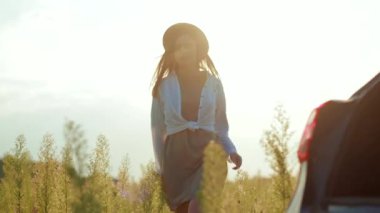 Güneş ışığında, şapkalı genç bir kadın arabasının yanında yürüyor. Gün batımının tadını çıkarıyor. Yüzeysel bir yüz ifadesi ile.