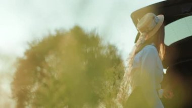 Şapkalı güzel genç bir kadın arabasının yanına bakar ve gün batımını seyreder.