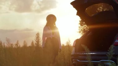 Şapkalı mutlu genç bir kadın akıllı telefon kullanıyor güneş ışığında şık bir arabanın bagajında oturuyor cep telefonu özgürlüğü nakliye aracı kız macera teknolojisinin tadını çıkarıyor yavaş çekim