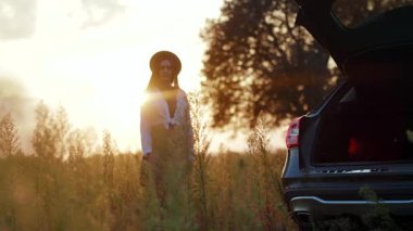 Güneş ışığında, şapkalı genç bir kadın arabasının yanında yürüyor. Gün batımının tadını çıkarıyor. Yüzeysel bir yüz ifadesi ile.