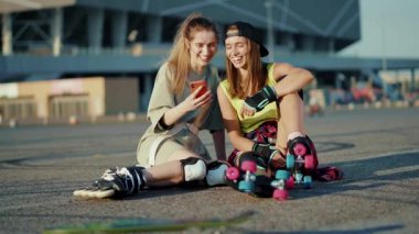 Patenlerle yerde oturan gülümseyen genç kadınlar yaz aylarında telefon kullanır. Ayaklar kayar, ağır çekimde hareket eder.