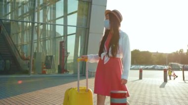 Korumalı maskeli bir kadın havaalanında tramvay bagajlı akıllı telefon kullanıyor. Corona virüsü salgını, yeni normal sosyal mesafe, havaalanı terminalinin dışında bekleyen kadın.