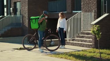 Genç adam kuryesini güvenlik kaskı ve tıbbi maskeyle kapatın. Müşteriye bisiklet çekici bir kadınla kese kağıdıyla yemek dağıtın. Dışarıda durun.