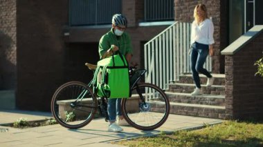 Güvenlik kaskı ve sağlık maskesi takmış kurye müşteriye bisiklet çekici kadın eşliğinde kese kağıdıyla yemek getiriyor. Dışarıda durup Coronavirus 'un daha az yavaş çekim teması kurmasını söyle.