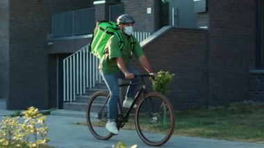 Dışarıda güvenlik kaskı ve tıbbi maske takmış genç bir kurye bisiklet siparişi Coronavirus ile yemek getiriyor.