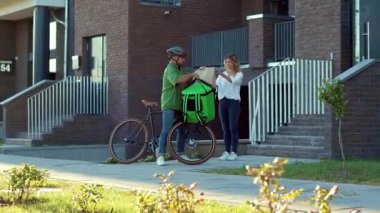 Güneş ışığı kuryesi kasklı bir kurye müşteriye kese kağıdıyla yemek getiriyor. Bisikletli çekici bir kadın dışarıda bekliyor. Yavaş çekim sırasını kapatın.