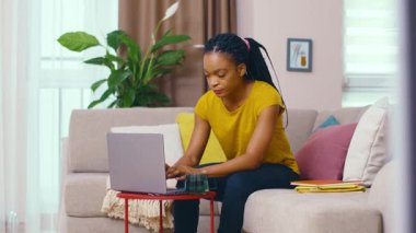 Serbest çalışan siyahi genç iş kadını internette iş yapmak için dizüstü bilgisayar kullanıyor oturma odasında oturarak uzak projeler yapıyor..
