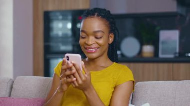 Neşeli duygusal siyahi kadın akıllı telefon kullanıyor sosyal medyada komik fotoğraflar çekiyor gülerek evde rahat bir hayat sürüyor. Teknoloji aygıtları.