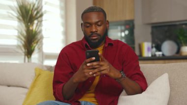 Afrika kökenli genç bir Amerikalı adam telefon kullanır. Etrafa bakar, evdeki kanepeye oturur. Sosyal medyada iletişim. Yakışıklı, ciddi bir adam. - İnternet. Yavaş çekim