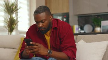 Afrika kökenli genç bir Amerikalı adam telefon kullanıyor. Evdeki kanepede oturuyor. Sosyal medyada iletişim kurmak iş uygulamasını gevşetiyor. Yakışıklı, ciddi bir adam. - İnternet. Yavaş çekim