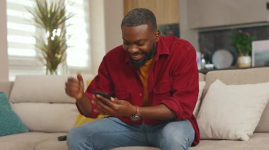 Mutlu Afrikalı genç Amerikalı adam telefon kullanıyor gülümsüyor iyi haberler okuyor evdeki kanepede oturuyor. Sosyal medyada iletişim kurmak iş uygulamasını gevşetiyor. Yakışıklı adammış. - İnternet. Yavaş çekim