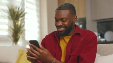 Portre mutlu genç Afrikalı Amerikalı adam telefon kullanıyor gülümsüyor iyi haberler okuyor evdeki kanepede otur. Sosyal medyada iletişim kurmak iş uygulamasını gevşetiyor. Yakışıklı adammış. - İnternet. Yavaş çekim