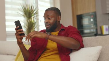 Mutlu, Afro-Amerikalı bir adam telefonuyla gülümsüyor. İyi haberler okuyor. Evdeki kanepede oturuyor. Sosyal medyada iletişim kurmak iş uygulamasını gevşetiyor. Yakışıklı adammış. - İnternet. Yavaş çekim