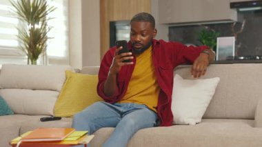 Gülümseyen mutlu Afro-Amerikalı adam telefon kullanıyor. İyi haberler okuyor. Evdeki kanepede otur. Sosyal medyada iletişim kurmak iş uygulamasını gevşetiyor. Yakışıklı adammış. - İnternet. Yavaş çekim
