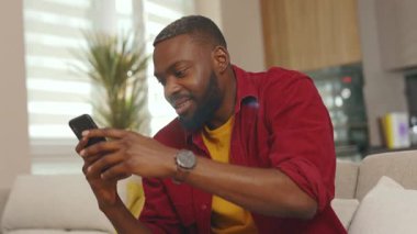 Şirin, mutlu, Afro-Amerikan bir adam telefon kullanıyor gülümsüyor, iyi haberler okuyor evdeki kanepede oturuyor. Sosyal medyada iletişim kurmak iş uygulamasını gevşetiyor. Yakışıklı adammış. - İnternet. Yavaş çekim