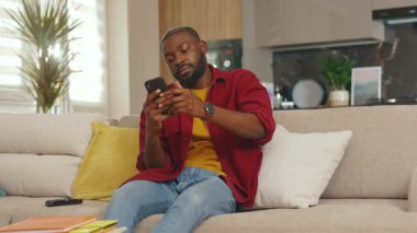 Ciddi bir Afrikalı genç Amerikalı evdeki kanepede telefon kullanıyor. Sosyal medyada iletişim kurmak iş uygulamasını gevşetiyor. Yakışıklı adammış. - İnternet. Yavaş çekim