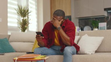 Yorgun, Afro-Amerikan bir adam telefon kullanıyor. Evdeki kanepede oturuyor. Sosyal medyada iletişim kurmak iş uygulamasını gevşetiyor. Yakışıklı adammış. - İnternet. Yavaş çekim