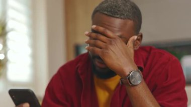 Portre yorgun Afrikalı Amerikalı genç adam telefon kullanıyor. Evdeki kanepede otur. Sosyal medyada iletişim kurmak iş uygulamasını gevşetiyor. Yakışıklı adammış. - İnternet. Yavaş çekim