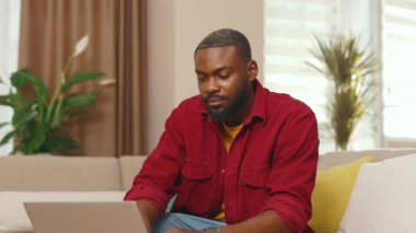 Dizüstü bilgisayar kullanan gülümseyen Afro-Amerikan genç adam evde çalışırken kanepeye oturmuş kameraya bakıyor. Teknoloji düşünen siyah daire bilgisayarı kapalı mekanda. Yavaş çekim