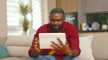 Gülümseyen Afrikalı Amerikalı genç adam evdeki dijital tablet bilgisayarı kullanarak koltukta oturuyor. Yakışıklı adammış. Teknoloji, siyah dairenin kapalı olduğunu düşünüyor. Yavaş çekim