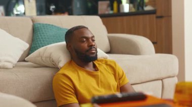 Rahatlamış Afro-Amerikan genç bir adam dinleniyor. Kanepenin yanında oturuyor, gözleri kapalı. Olumlu düşünceli meditasyon. Memnuniyet gülümsemesi. Denge uyumu. Yavaş çekim