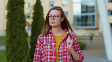 Gözlüklü ve çantalı başarılı genç kız üniversite kampüs binasının yakınındaki sokakta kameraya bakıyor. Eğitim. Üretim.