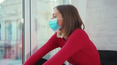 Üzgün genç kadın pencere pervazında oturur. Tıbbi yüz maskesi çıkartır. Koronavirüs kendini izole eder.