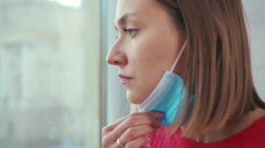 Mutsuz genç bir kadın pencere pervazına oturmuş tıbbi yüz maskesi takıyor koronavirüs kendini tecrit depresyon penceresi sıkıcı ağır çekim depresyon penceresi