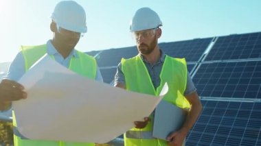 Güneş ışığında güneş çiftliğinde proje planına sahip iki mühendis ayakta durmuş güneşli pillerin kurulumunu tartışıyorlar. Elektrik yavaş çekim.