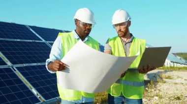 Erkek mühendisler güneş çiftliğinde dizüstü bilgisayar ve proje planı kullanıyorlar. Güneşli bataryaların kurulumunu tartışıyorlar. Afrika kökenli Amerikan elektriği yavaş çekim.