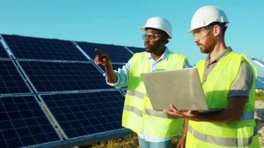 İki adam güneş çiftliğinde dizüstü bilgisayar ve proje planı kullanıyor. Güneşli bataryaların kurulumunu tartışıyorlar. Afrikalı Amerikan elektriği yavaş çekim.