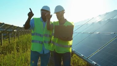 Güneş ışığında iki adam güneş çiftliğinde dizüstü bilgisayar kullanıyor. Güneşli bataryaların kurulumunu tartışıyorlar. Afrika kökenli Amerikan elektriği yavaş çekim.