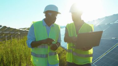 Ağır çekimde iki adam güneş çiftliğinde dizüstü bilgisayar kullanıyor. Özel üniformalı yürüyorlar. Güneşli bataryaların kurulumunu tartışıyorlar.